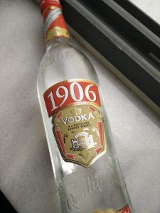 A 700ml clear glass bottle of Polmos Lublins 1906 Vodka sits on a white surface near a window, its red and gold label displayed. Quadruple distilled for exceptional smoothness, the bottle tilts slightly to the right.